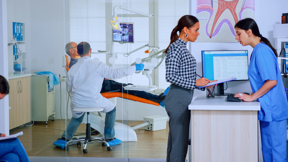 Dental practice staff reviewing records at the front desk while a dentist treats a patient